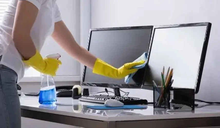 Person wearing yellow gloves cleaning computer monitors on an office desk, illustrating professional commercial cleaning services