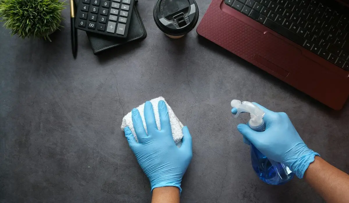 Hands wearing blue gloves cleaning a desk with a spray bottle and cloth as part of eco-friendly commercial cleaning.