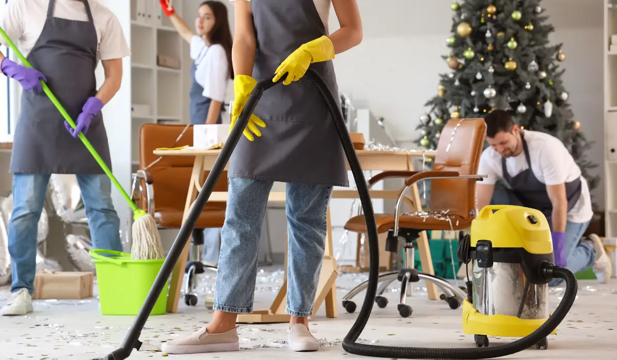 Cleaning crew performing holiday cleaning rush tasks in decorated office after a staff event with visible clutter.